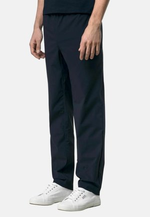 Uomo con pantaloni neri a gamba dritta e sneaker bianche basse, in piedi davanti a uno sfondo semplice.