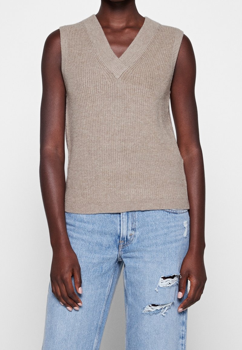 Pull sans manches beige en maille avec un col en V et une texture côtelée. Associé à un jean bleu clair déchiré avec des déchirures au niveau du genou.