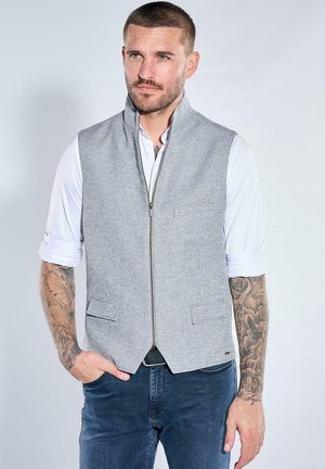 Mann mit Tattoos, der eine hellgraue Reißverschlussweste über einem weißen hochgekrempelten Hemd und dunkelblaue Jeans trägt, stehend mit einer Hand in der Tasche.