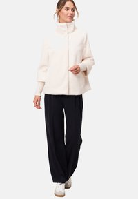 Veste en peluche de couleur claire avec un col montant, dotée d'une fermeture éclair à l'avant et de poignets élastiques, associée à un pantalon large noir et des baskets blanches.