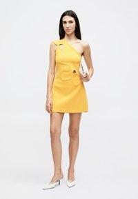 Femme debout portant une mini robe jaune à une épaule avec des découpes, tenant une pochette blanche et portant des talons pointus blancs.