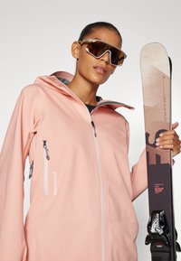 Svetle růžová lyžařská bunda s kapucí, předním zipem a bočními kapsami. Model drží lyži s geometrickým vzorem a vázáním. Tmavé sluneční brýle.