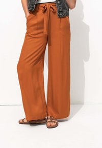 Pantalons larges en tissu orange brûlé avec une texture lisse, dotés d'une taille nouée, de poches latérales et d'un design léger.
