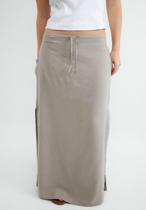 Kobieta nosząca długą, jasnoszarą spódnicę z wiązaniem w talii i bocznymi rozcięciami, połączona z białym krótkim topem bez rękawów.