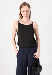 Melna satīna kamizola ar šaurām siksnām, apvienota ar jūras zila krāsas kroku biksēm. Modelis tur nelielu melnu rokassomu ar unikālu formu.