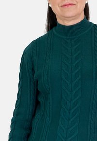 Suéter de punto en color teal con un cuello alto. Presenta patrones de punto trenzado y puños acanalados, mostrando un acabado texturizado.