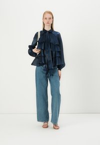Blouse en satin navy avec des couches froncées, col montant, manches longues, portée avec un jean large bleu clair et des sandales à lanières, tenant un sac.