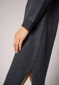 Sweter sukienka w kolorze węgorzowym z żebrowymi mankietami i ozdobami w postaci kamieni szlachetnych na rękawie. Posiada boczne rozcięcia oraz miękką, teksturowaną dzianinę.
