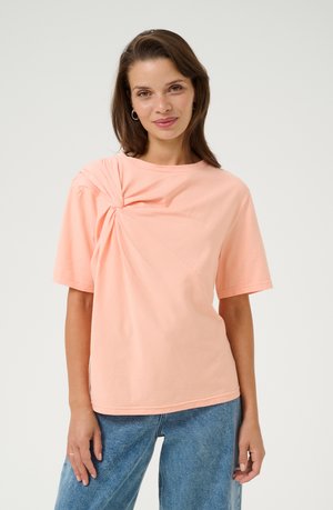 Peach-colored cotton t-shirt featuring a twisted knot detail on the left shoulder and short sleeves, paired with high-waisted blue jeans.