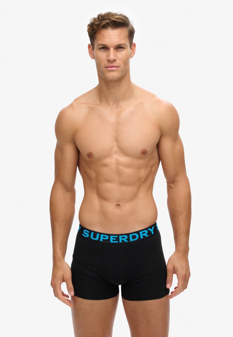 Caleçons boxers noirs avec une ceinture logo "SUPERDRY" bleue, texture lisse et design ajusté. Modèle présenté avec une silhouette musclée et sans chemise.