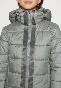 Šedá péřová bunda s předním zipem, která má dvouřadý šev a tmavě šedé vertikální pruhy na přední části. Součástí je nastavitelná kapuce.