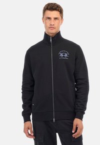 Fekete zipzáros pulóver magas nyakkal, hímzett logóval a mellkasán, bordázott mandzsetta és hem; az anyag lágy és texturált.