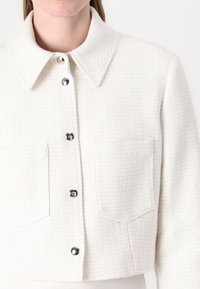 Veste blanche texturée avec un col, dotée de quatre boutons noirs et de deux poches avant. Design court et cropped.