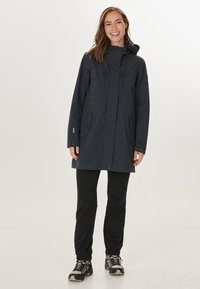 Marineblauwe waterdichte parka met capuchon, verstelbare manchetten, twee voorkakken en een rechte zoom. Gecombineerd met zwarte broek en wandelschoenen.