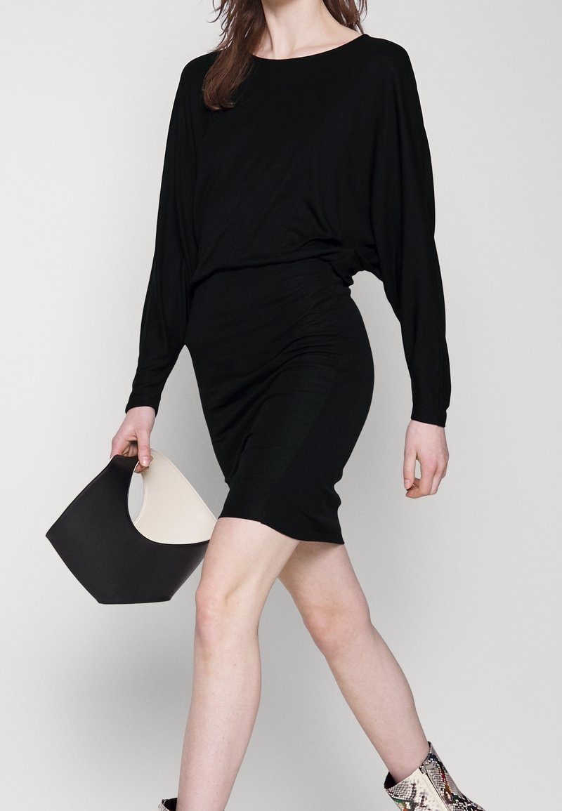 Czarna sukienka z długim rękawem o dopasowanym kroju, w połączeniu z czarną i kremową torebką, noszona z wzorzystymi botkami za kostkę.