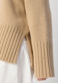 Beige gebreide trui met geribbelde manchetten en zijsplitten. De stof heeft een zachte textuur, met een losvallende pasvorm en lange mouwen.