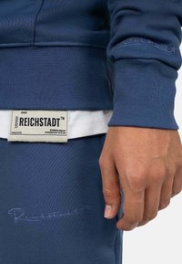 Felpa blu navy con polsini a coste, caratterizzata da un logo ricamato in azzurro e un'etichetta sull'orlo inferiore con testo del brand e istruzioni per la cura.