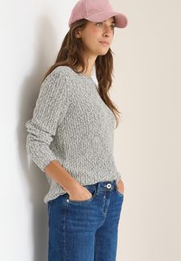 Un pull gris côtelé à manches longues, associé à un jean en denim bleu foncé. Une casquette en velours côtelé rose clair ajoute un élément de contraste.