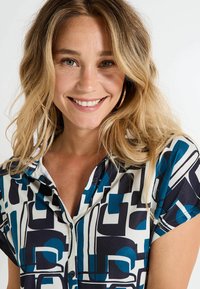 Femme souriante aux cheveux blonds portant des boucles d'oreilles en or et un haut à manches courtes avec un motif géométrique bleu, noir et blanc sur fond blanc.