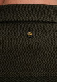 Tejido verde oscuro con un patrón de espiga, con una etiqueta negra que lleva el texto dorado "PME LEGEND".