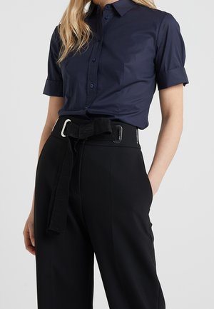Femme portant une chemise bleu marine à manches courtes boutonnée, rentrée dans un pantalon noir taille haute avec une large ceinture en tissu nouée en nœud.