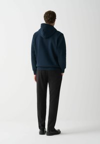 Tumed kapuutsiga sweatshirt, kus on ees tasku ja reguleeritav kapuuts, koos mustade pükste ja kingadega, näidates ühtlast kangasteksti.