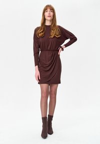 Kobieta o długich włosach, ubrana w dopasowaną brązową sukienkę z długim rękawem i pasujące botki do kostki, stoi z jedną ręką na biodrze na białym tle.