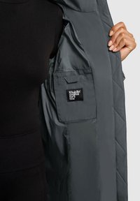 Grå quiltet jakke indvendig med et sort mærke; har en sidelomme og glat for. Ydersiden viser en uldet front.