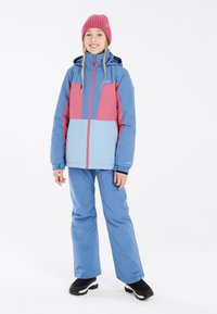 Schneejacke in Blau und Pink, mit einem Frontreißverschluss, verstellbarem Kapuze und passenden blauen Schneehosen. Getragen mit schwarzen Schneestiefeln.