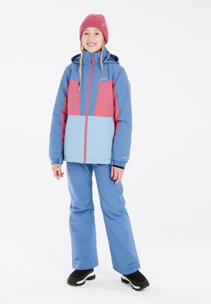 Giacca da neve blu e rosa, con zip frontale, cappuccio regolabile e pantaloni da neve blu coordinati. Indossata con stivali da neve neri.