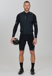 Schwarze Radweste mit Reißverschluss, schwarze lange Ärmel, schwarze Shorts, schwarze Socken und schwarze Radschuhe. Hält einen schwarzen Helm.