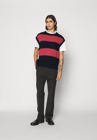 Gilet en tricot avec des rayures rouges et bleu marine sur un t-shirt blanc ; associé à un pantalon sombre et des chaussures noires, avec une coupe décontractée.