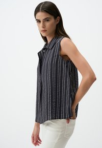 Canotta in blu scuro con righe verticali bianche, dotata di collo a camicia e tasche laterali. Tessuto testurizzato con vestibilità rilassata.