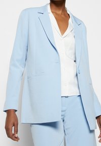Blazer de color azul claro con un diseño ajustado, que presenta solapas de muesca, cierre de un solo botón y dos bolsillos frontales, hecho de una tela suave.