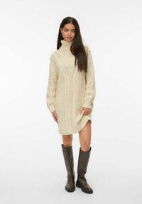 Kvinna i en gräddfärgad kabelstickad turtleneck klänning och mörkbruna knähöga stövlar, stående mot en vit bakgrund.