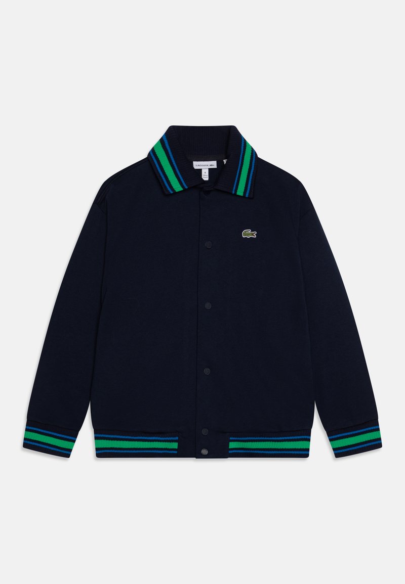 Giacca a maglia blu navy con colletto a coste e strisce blu e verdi, chiusura con bottoni. Logo Lacoste sul petto sinistro.