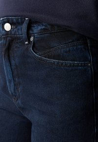 Close-up van donkerblauwe denim jeans met zichtbare stiksels, een metalen knoop en details van de zakken onder een marineblauw shirt.