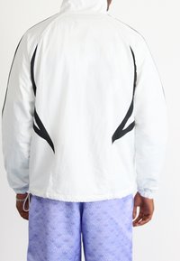 Vit vindjacka med svarta accenter och böjda sidsömmar, med hög krage och elastiska ärmslut, tillsammans med lila mönstrade byxor.