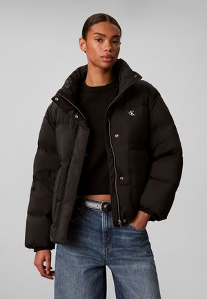 Jeune personne portant une doudoune noire par-dessus un pull noir et un jean bleu, avec une main dans la poche et une expression neutre.