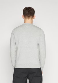 Uomo di spalle che indossa un maglione grigio chiaro a maniche lunghe e pantaloni neri su uno sfondo bianco semplice.