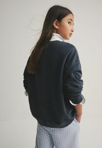 Marineblaues Sweatshirt mit überschnittenen Schultern, gerippten Bündchen und lockerer Passform, über einem weißen Hemd mit Kragen getragen. Hellblaue, vertikal gestreifte Hose.