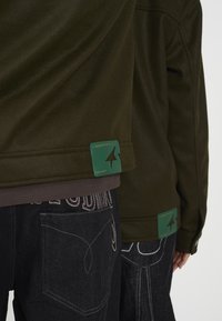 Grønn fleecejakke med teksturert overflate og en grønn etikett med en pil. Sorte jeans med kontrasterende sømmer og lomme detaljer.