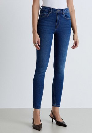 Donna che indossa jeans skinny blu scuro a vita alta e tacchi neri con punta affilata, in piedi davanti a uno sfondo chiaro e semplice.