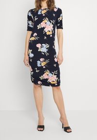 Nainen pukeutuneena tummansiniseen polvipituiseen kukkamekkoon, jossa on pinkkejä, sinisiä ja keltaisia kukkia, yhdessä mustien avokkaiden kanssa.