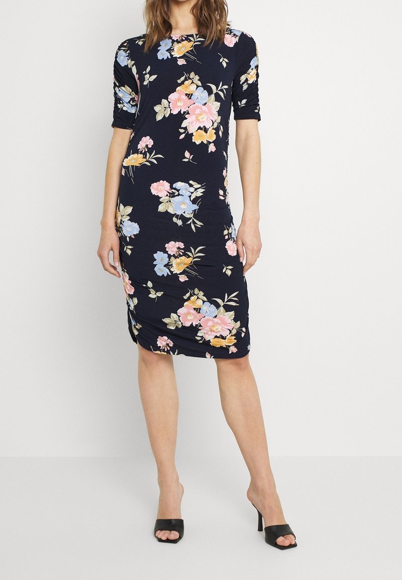 Nainen pukeutuneena tummansiniseen polvipituiseen kukkamekkoon, jossa on pinkkejä, sinisiä ja keltaisia kukkia, yhdessä mustien avokkaiden kanssa.