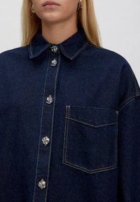 Donkerblauw spijkerblouse met een puntkraag, zilveren decoratieve knopen en een borstzak. Kenmerkt zichtbare stikseldetails.