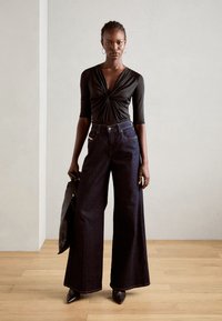 Svart langermet topp med vridd front, kombinert med høytlivede, vide mørk denim jeans. Modellen holder en svart veske og har på seg spisse hæler.