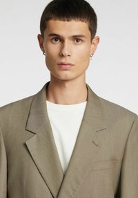 Bež srajca s širokimi reverji, enim žepom in subtilno teksturo. Nošena čez belo srajco. Minimalističen dizajn s čistimi linijami.