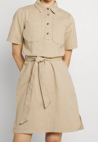 Robe beige à manches courtes avec boutons sur le devant, poches poitrine et ceinture en tissu nouée à la taille.