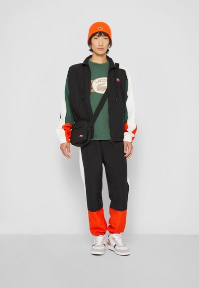 Gilet noir zippé avec des accents blancs et orange sur une chemise verte. Pantalon noir avec des manchettes orange. Bonnet en tricot orange ; petit sac noir.